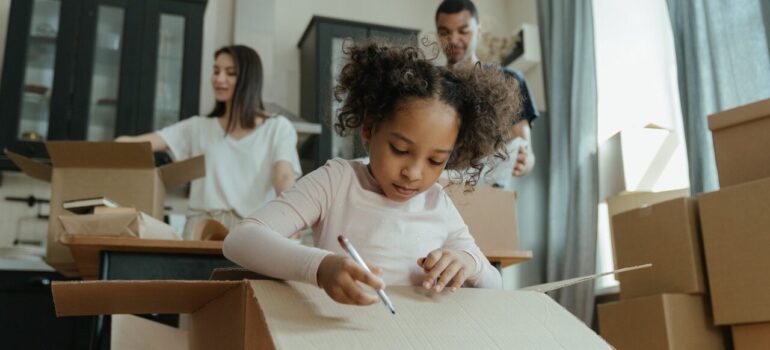 a small girl drawing on a cardboard box while her parents pack in the background