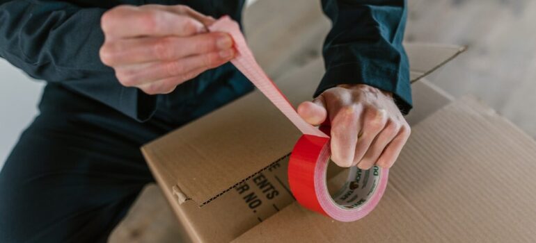 a moving crew personnel stretching a red tape to tape the cardboard box