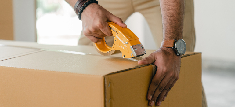 A man sealing shut a moving box containing pieces of furniture.