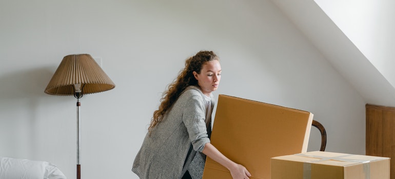 Woman lifting heavy cardboard box while standing near floor lamp