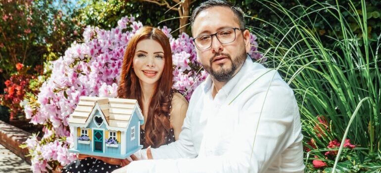 Man and woman surrounded by flowers holding a small house model happy after finding a perfect home in Maryland