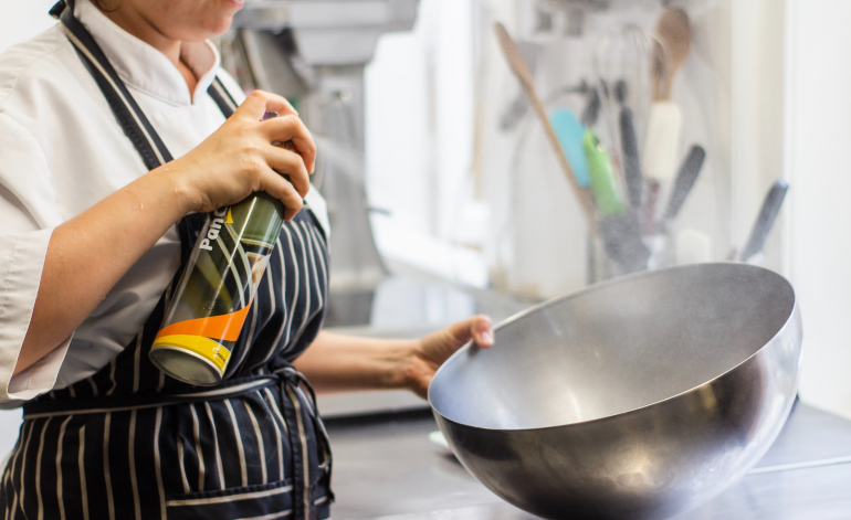 A woman aplying some sort of spray inside a metal bowl in a kitchen.