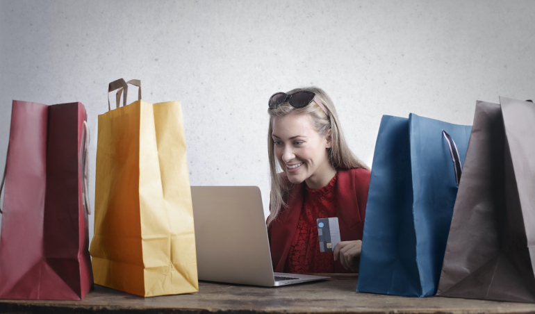A woman smiling while looking at a laptop next to several shopping bags. 