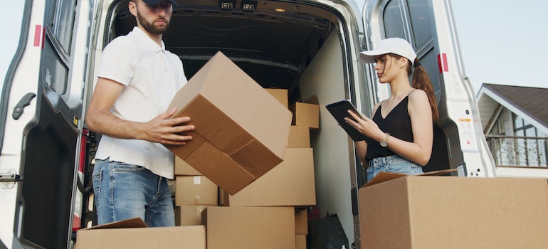 A woman and a man handling moving boxes in front of a van.