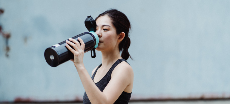 a woman in fitness clothes drinking water from a sports water bottle after reading the Alexandria's guide to outdoor fitness