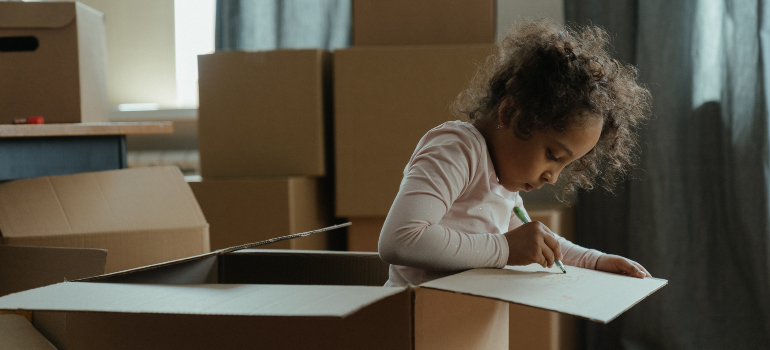 a little girl labeling a cardboard box while standing in it before moving from Ashburn to Frederick