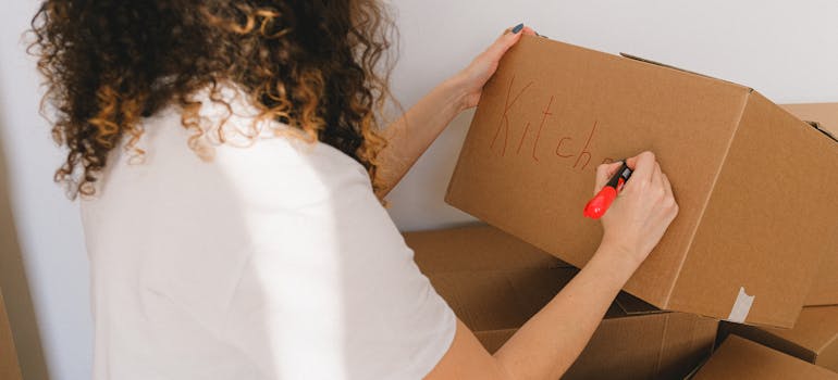 a woman writing the word "kitchen" on a cardboard box