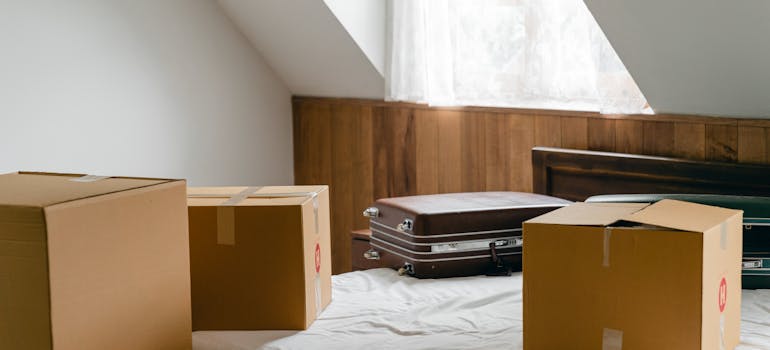 three cardboard boxes and a suitcase on a bed in a room with lots of sunlight