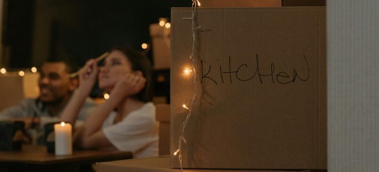 A couple sitting among moving boxes, with a 'kitchen' labeled box in the foreground and soft lighting creating a warm, intimate atmosphere.