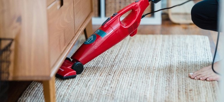 A person vacuuming under a cabinet with a red vacuum cleaner on a woven rug, ensuring the space is spotless.