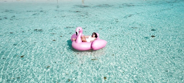 A woman relaxes on a pink flamingo float in crystal-clear tropical water—Book your summer move to total bliss.