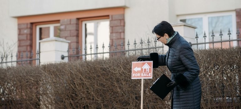 Woman in a black coat installing a "For Sale" sign outside a residential property in a quiet neighborhood.