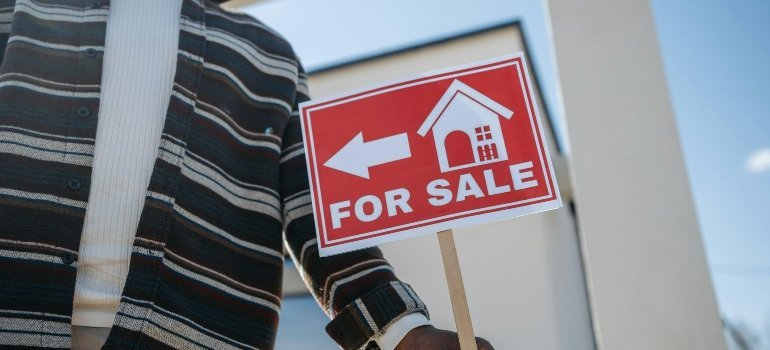 Person in striped sweater holding a "For Sale" sign in front of a contemporary home with blue sky backdrop.