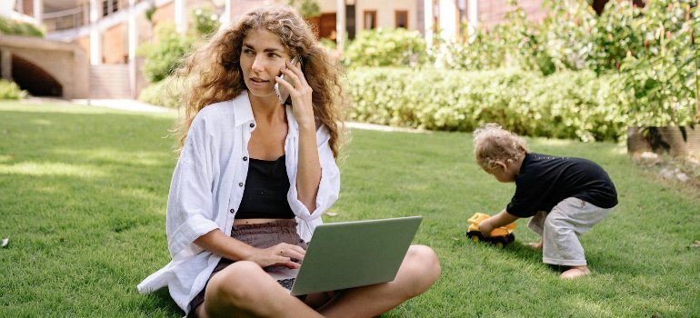 Mother balancing remote work and childcare outdoors while her toddler plays, reflecting the lifestyle changes after moving for a job with a family.