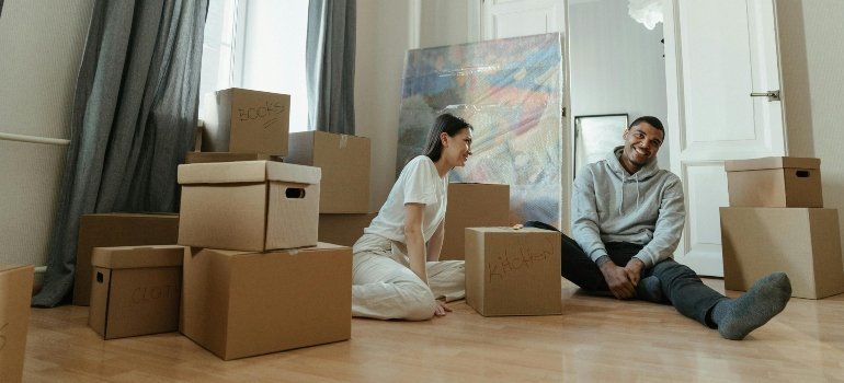 A smiling couple seated on the floor, surrounded by moving boxes labeled "Kitchen" and "Books."