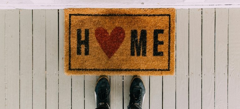 A doormat with the word “HOME” featuring a heart symbol, seen from above with feet in frame — highlighting What Makes Clinton MD a Great Place to Live.