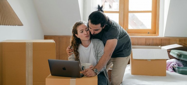 Young couple working together on a laptop, surrounded by moving boxes in their new bedroom.