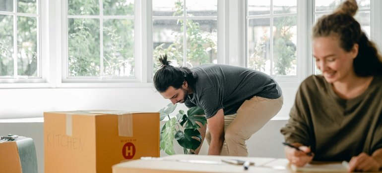 A man lifting a box labeled “Kitchen” while a woman nearby writes notes during moving from Baltimore to another Maryland city.