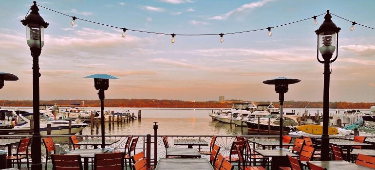 Empty tables and string lights at a cozy waterfront restaurant with docked boats in the background.