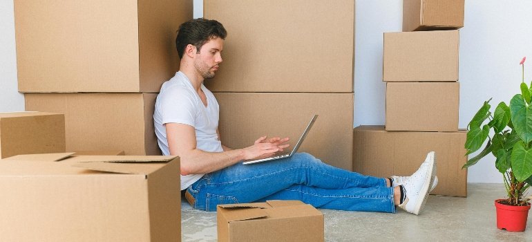 A man sits on the floor with his laptop, surrounded by moving boxes while organizing his relocation.