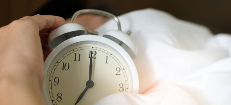 Close-up of a hand reaching for a white alarm clock resting on a bed with soft white sheets, signaling morning or wake-up time.