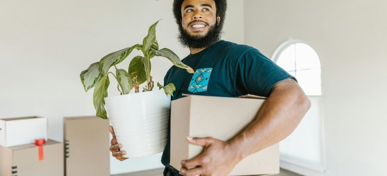Smiling man carrying a large houseplant and a cardboard box while moving into a bright, unfurnished space.