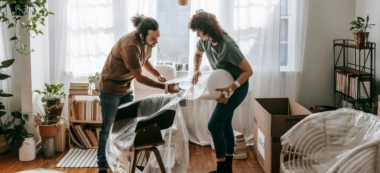 Couple carefully wrapping a chair in bubble wrap, as if preparing it for a fashion show.