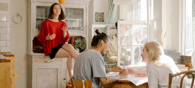 Three people at a kitchen table review papers while a woman in a red sweater perches nearby tossing an orange.