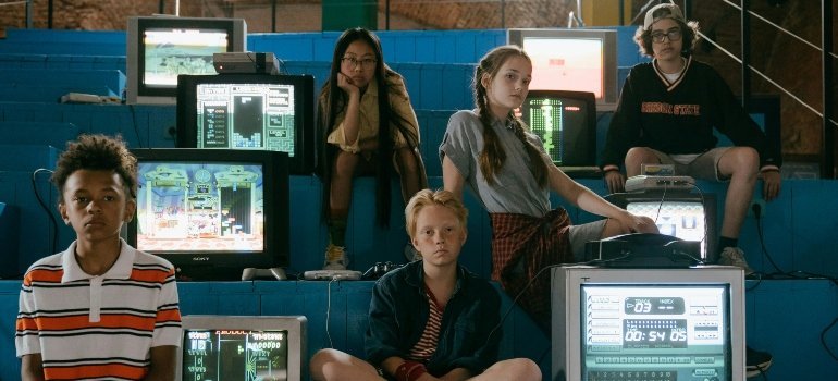 Group of teens hang out among retro CRT monitors in a rec room, relaxed and thoughtful.