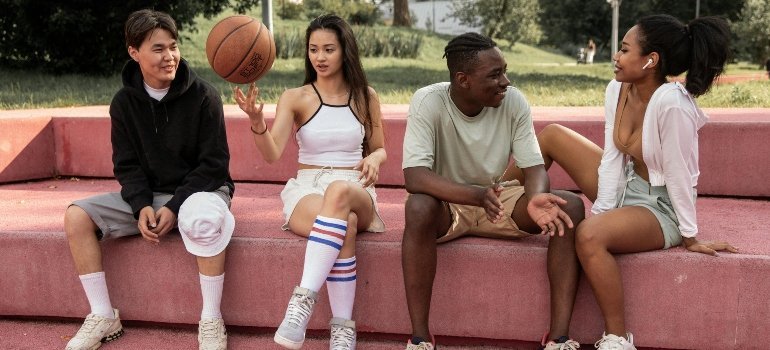 Four teens sit on outdoor steps by a basketball court, talking and smiling.