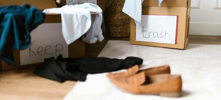 Cardboard boxes labeled Keep and Trash with clothes and shoes scattered on a rug during packing.