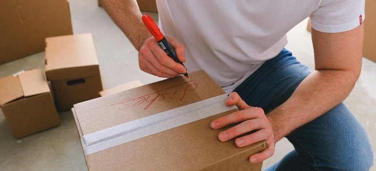 Person writing 'dishes' on a cardboard moving box with a red marker.