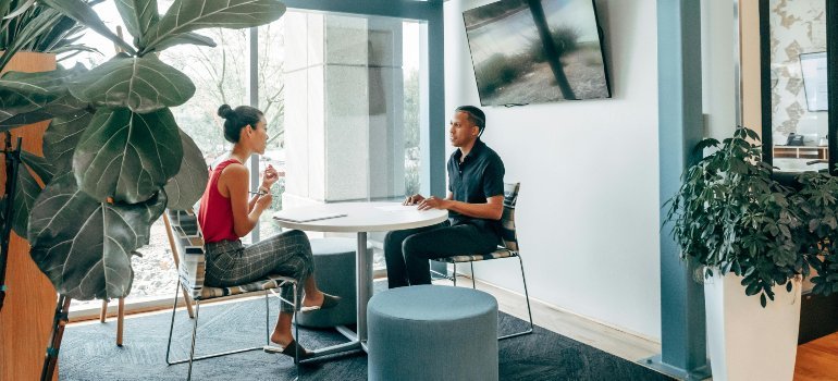 Two coworkers have a discussion at a small round table in a modern office space with large windows and indoor plants.