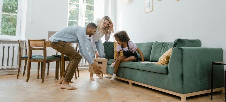 Parents lifting a moving box while their child sits on the sofa, smiling.