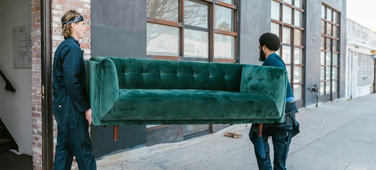 Two movers carrying a heavy green couch outdoors through a tight passage.