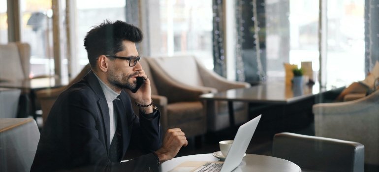 A man in a suit sitting at a table with a laptop and talking on the phone about choosing between hourly movers and flat-rate movers