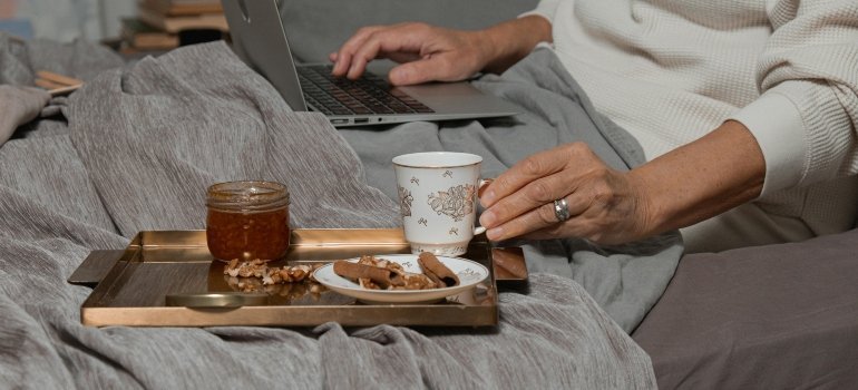 A person holding a coffee cup and using a laptop in bed to research how to live comfortably in Maryland.
