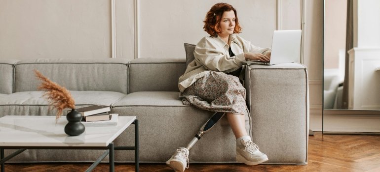 A woman with a prosthetic leg sitting on a sofa using her laptop in a bright living room.