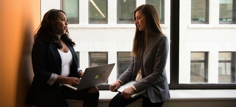 Two coworkers discussing ideas while working on a laptop near a window.