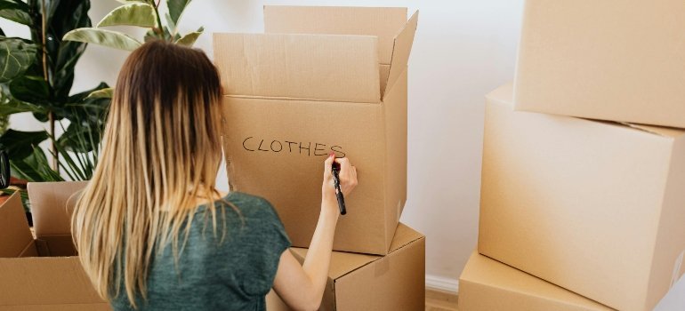Woman writing “clothes” on a moving box while organizing items for transport.