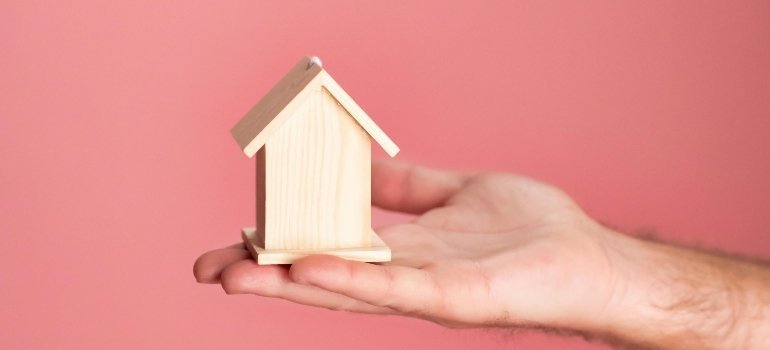 A small wooden model house resting in an open hand against a pink background, symbolizing a fresh housing opportunity.