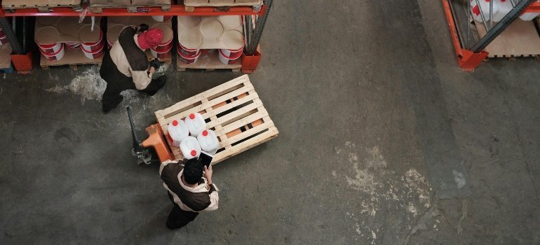 Two warehouse workers transport plastic containers on a pallet inside a storage facility.