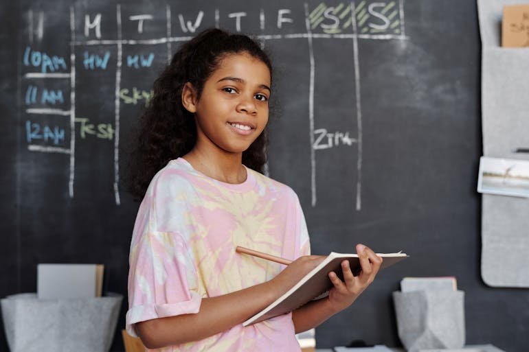 A teenage girl with a notebook and pencil standing in front of a blackboard calendar.