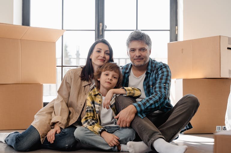 Family of three sitting on the floor surrounded by moving boxes in a bright room with large windows.