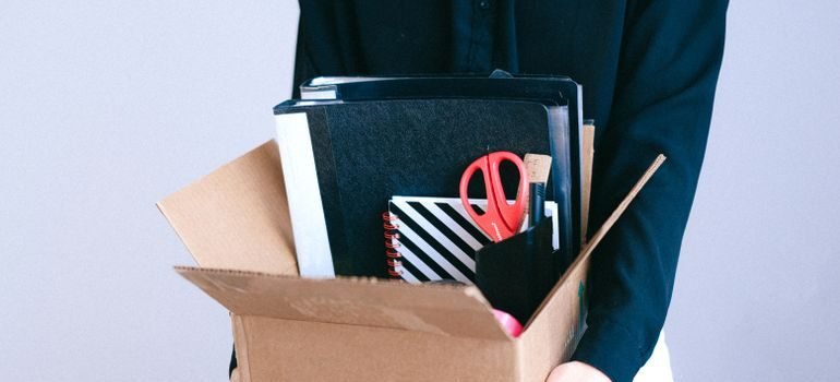 A person holding an open moving box packed with folders, notebooks, and scissors